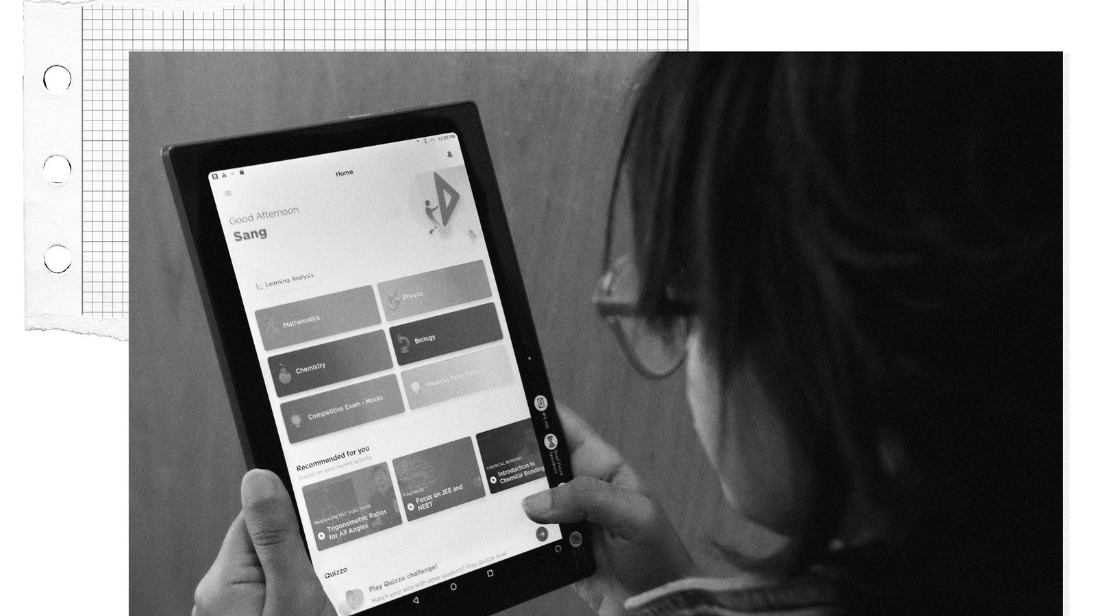 A person holding a tablet displaying a learning app interface with subjects like Mathematics, Physics, Chemistry, and Biology, while sitting against a wooden background. The screen also includes sections for recommendations and a quiz challenge.