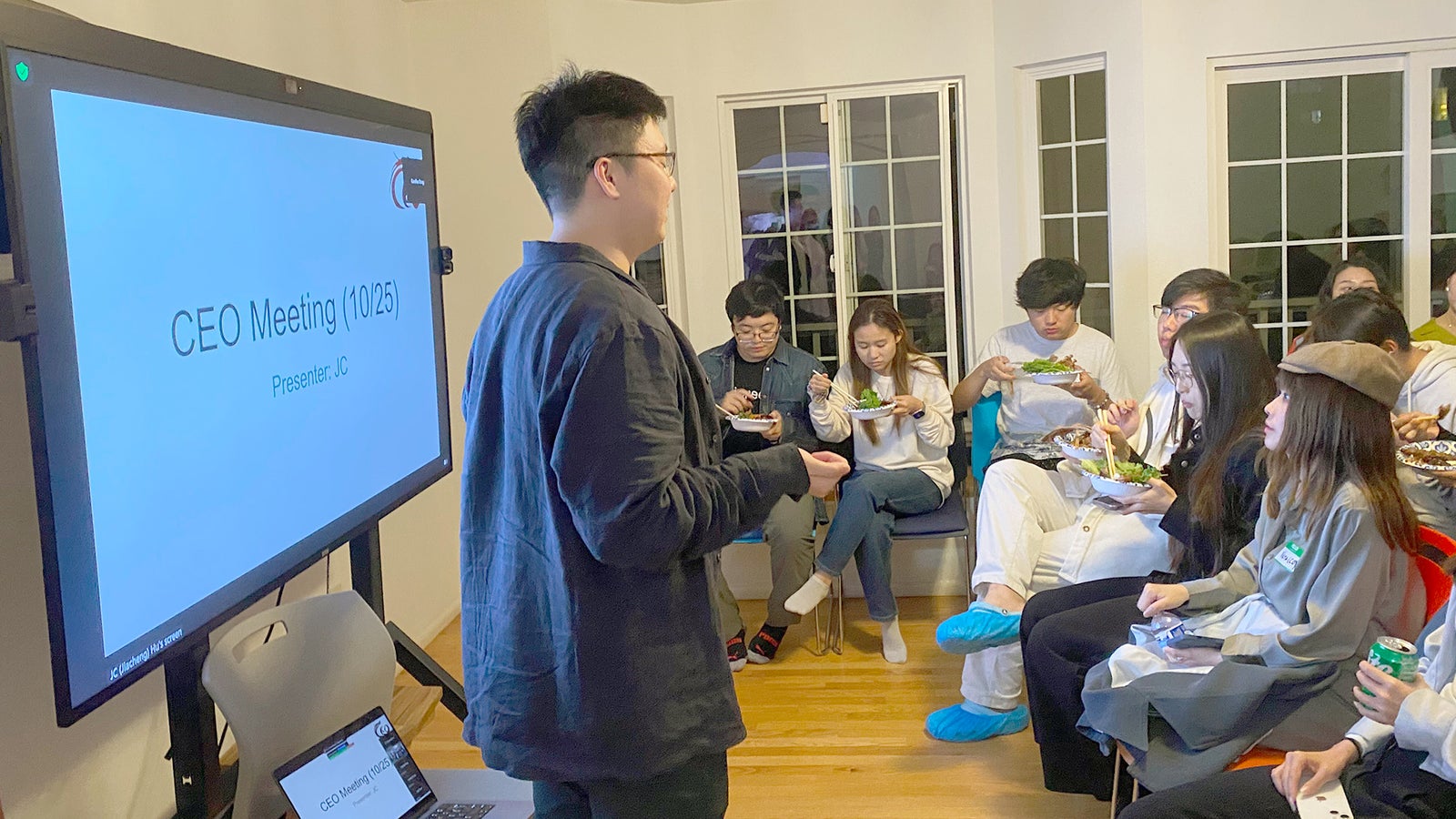 A speaker stands in front of a presentation screen titled "CEO Meeting," addressing a group of people seated and eating.