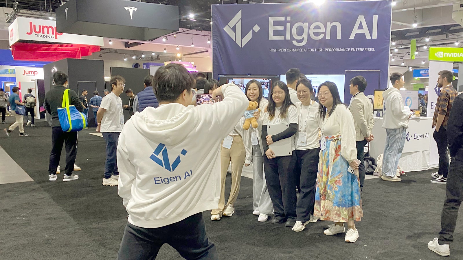 A group of people in white Eigen AI hoodies pose for a photo at a technology event, with a large Eigen AI banner in the background.