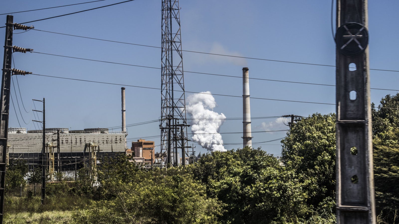 An industrial landscape with tall smokestacks releasing white smoke, surrounded by green foliage and power lines against a clear blue sky.