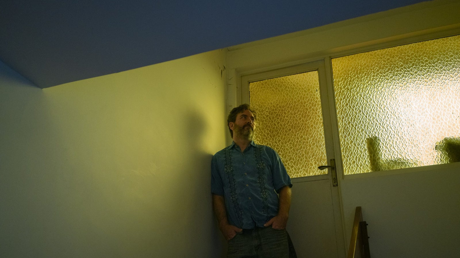 A portrait of Omar Alfonzo leaning against a wall taken in front of a door with frosted glass, through which a strong yellow-hue is is cast on Alfonzo.