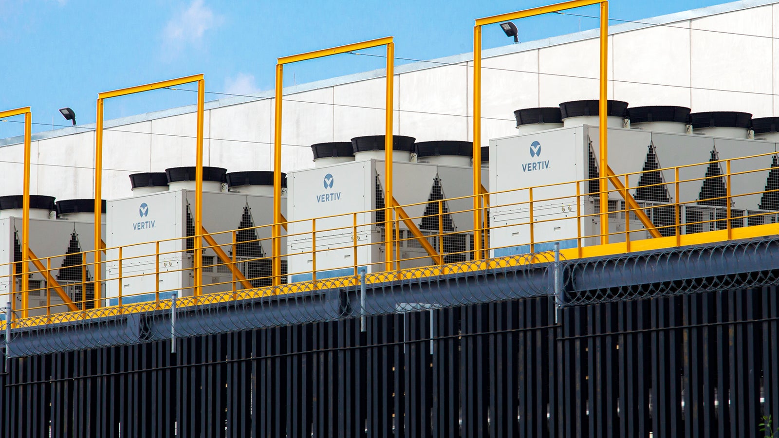 A row of large cooling units bearing the 'Vertiv' logo, positioned on an elevated platform with a yellow railing, against a backdrop of a white building and blue sky.