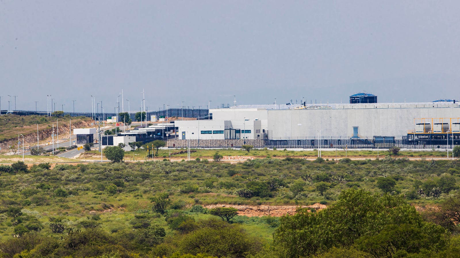 A large modern industrial facility situated in a green landscape, with access roads, security fencing, and wind turbines in the vicinity.