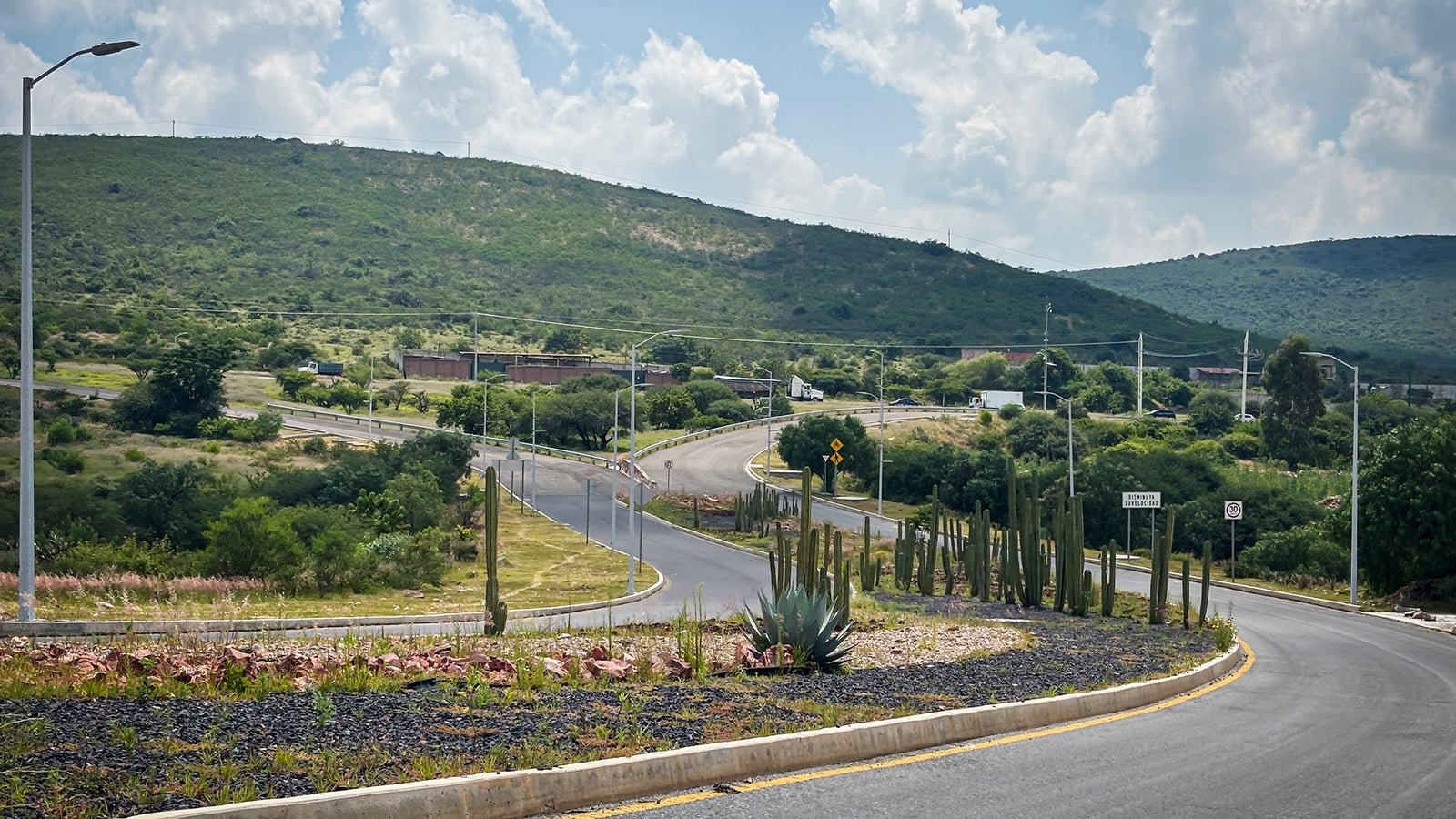 A winding road curves through a hilly landscape, bordered by cacti and sparse vegetation under a partly cloudy sky.
