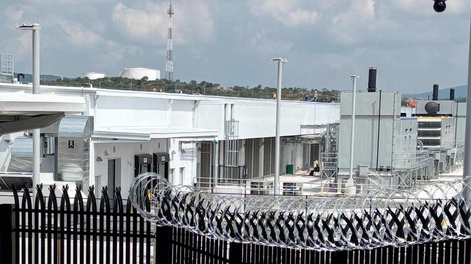 A fenced facility featuring barbed wire in the foreground, with industrial buildings and equipment in the background under a cloudy sky. A telecommunications tower and two large tanks are visible in the distance.