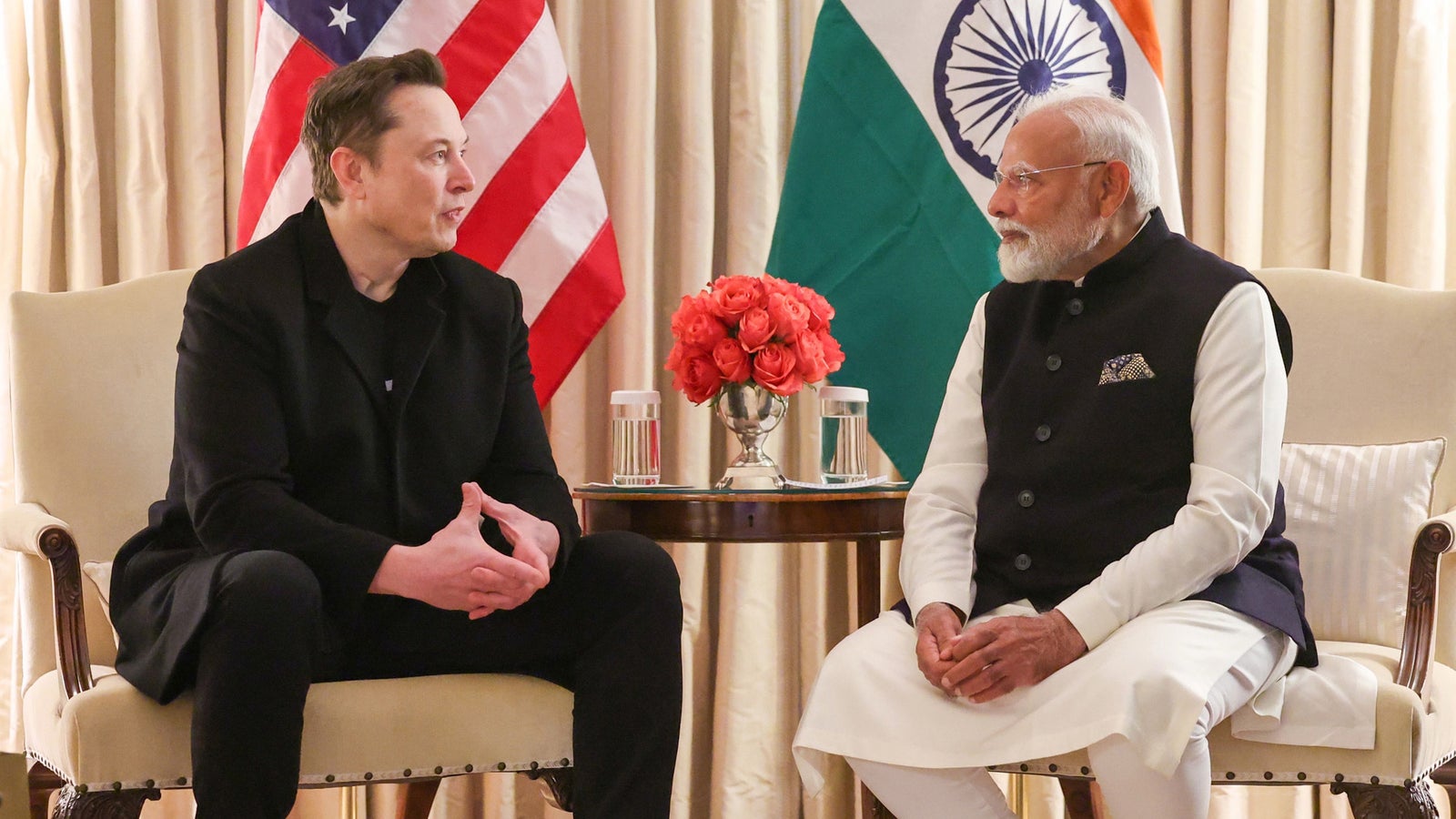 Two men seated face-to-face in a formal meeting, with the flags of the United States and India in the background. One man wears a black coat, while the other is in traditional Indian attire with a white kurta and a navy waistcoat. A vase of peach roses and water glasses are positioned on a small table between them.
