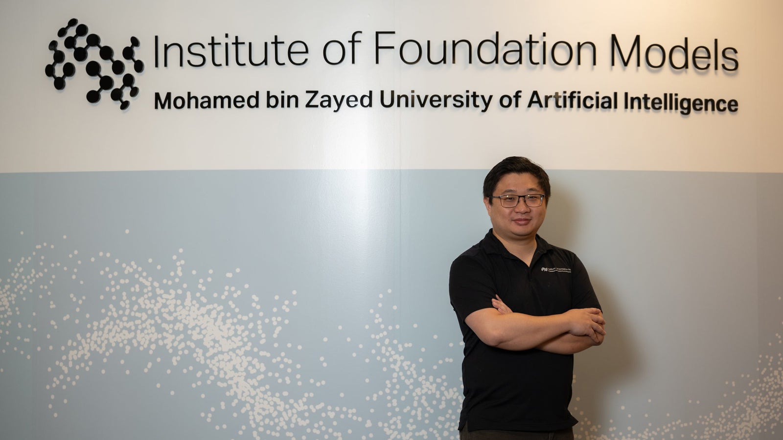 A person stands with arms crossed in front of a wall featuring the logo of the Institute of Foundation Models at Mohamed bin Zayed University.