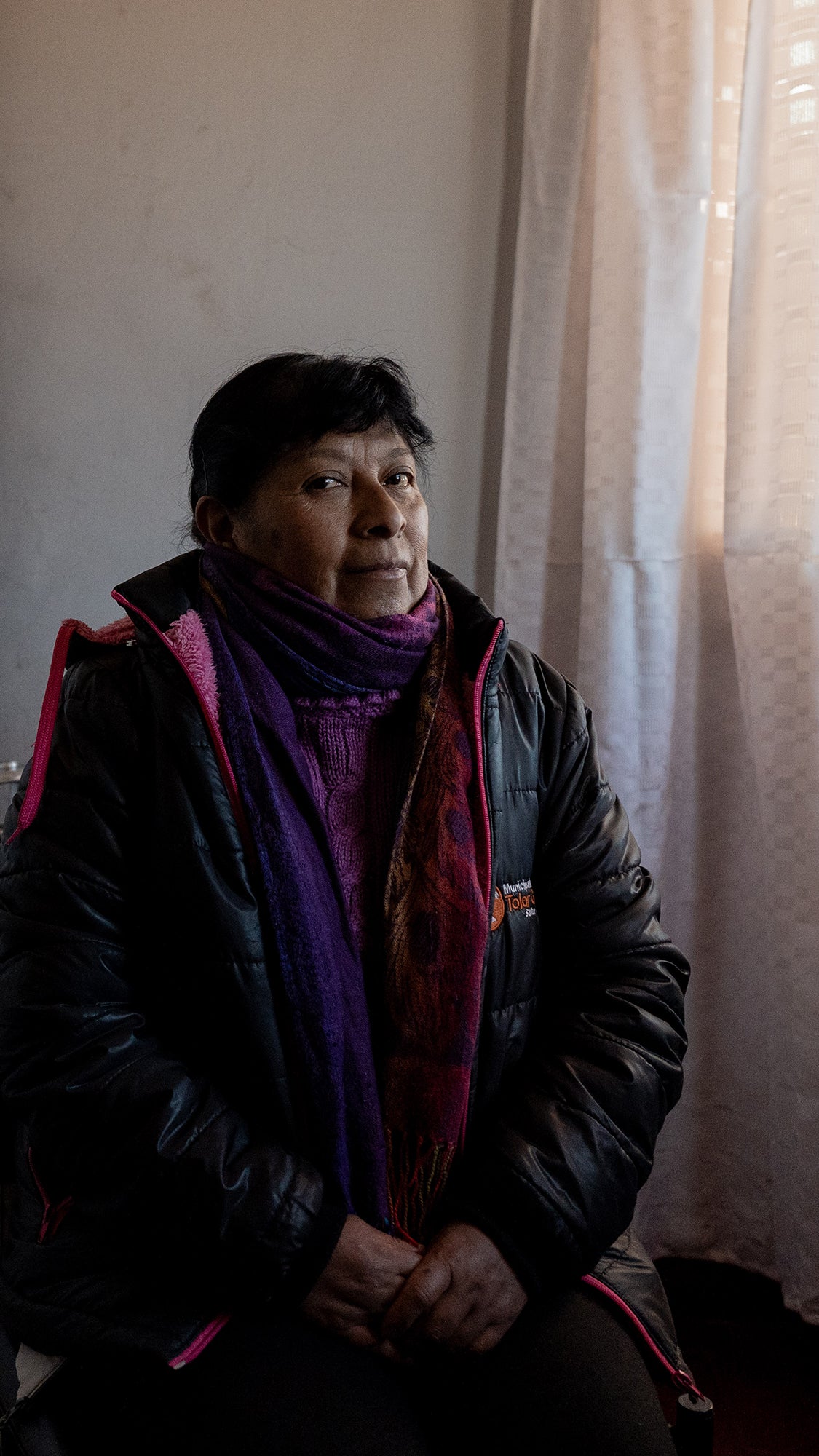 A woman wearing a black puffer jacket and colorful scarf sits against a wall with light curtains.