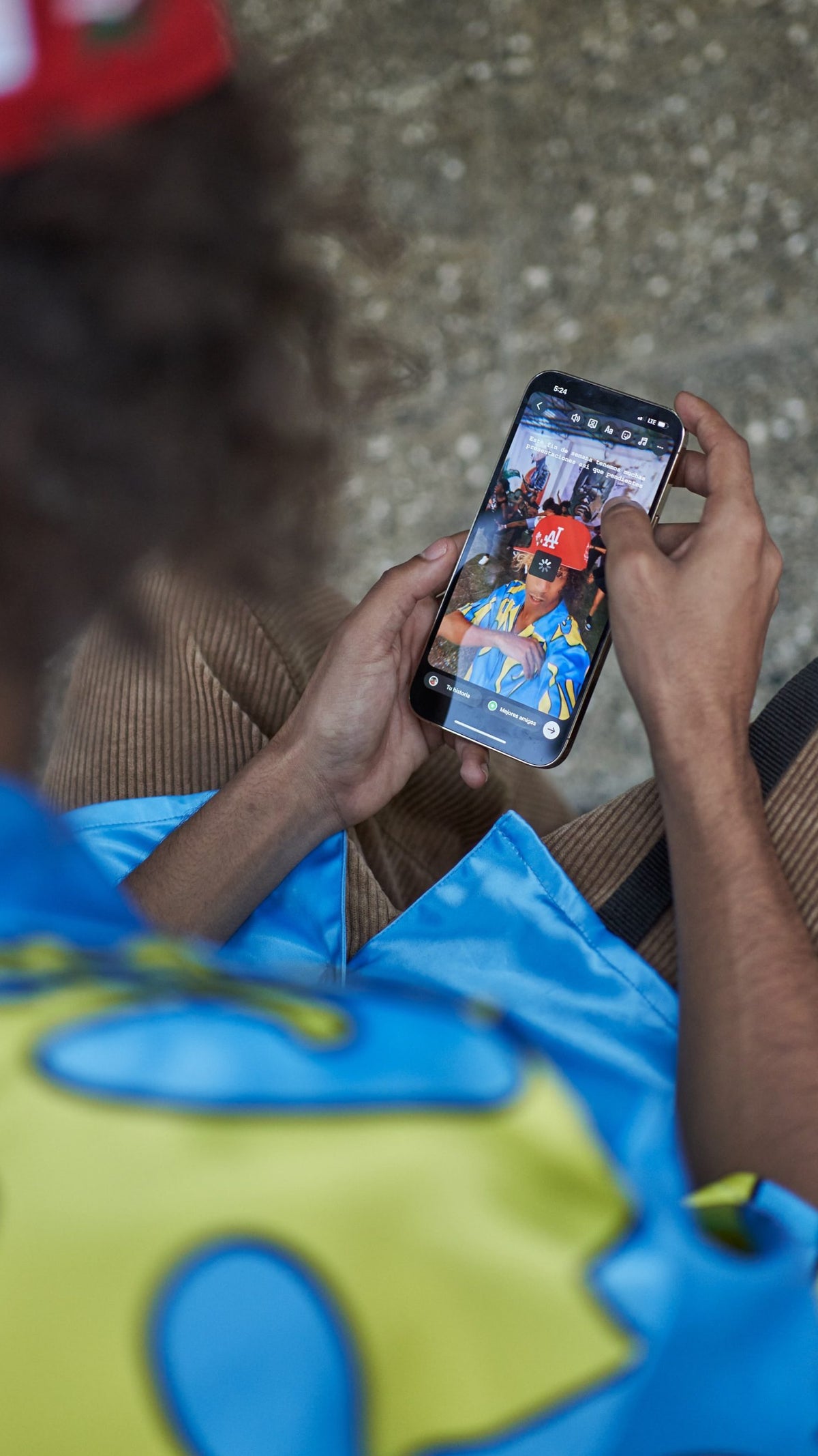 A photograph showing a person holding a mobile phone in their hands while seated. On the phone's screen is a loading circle over an image of the same individual inside an Instagram edit screen.