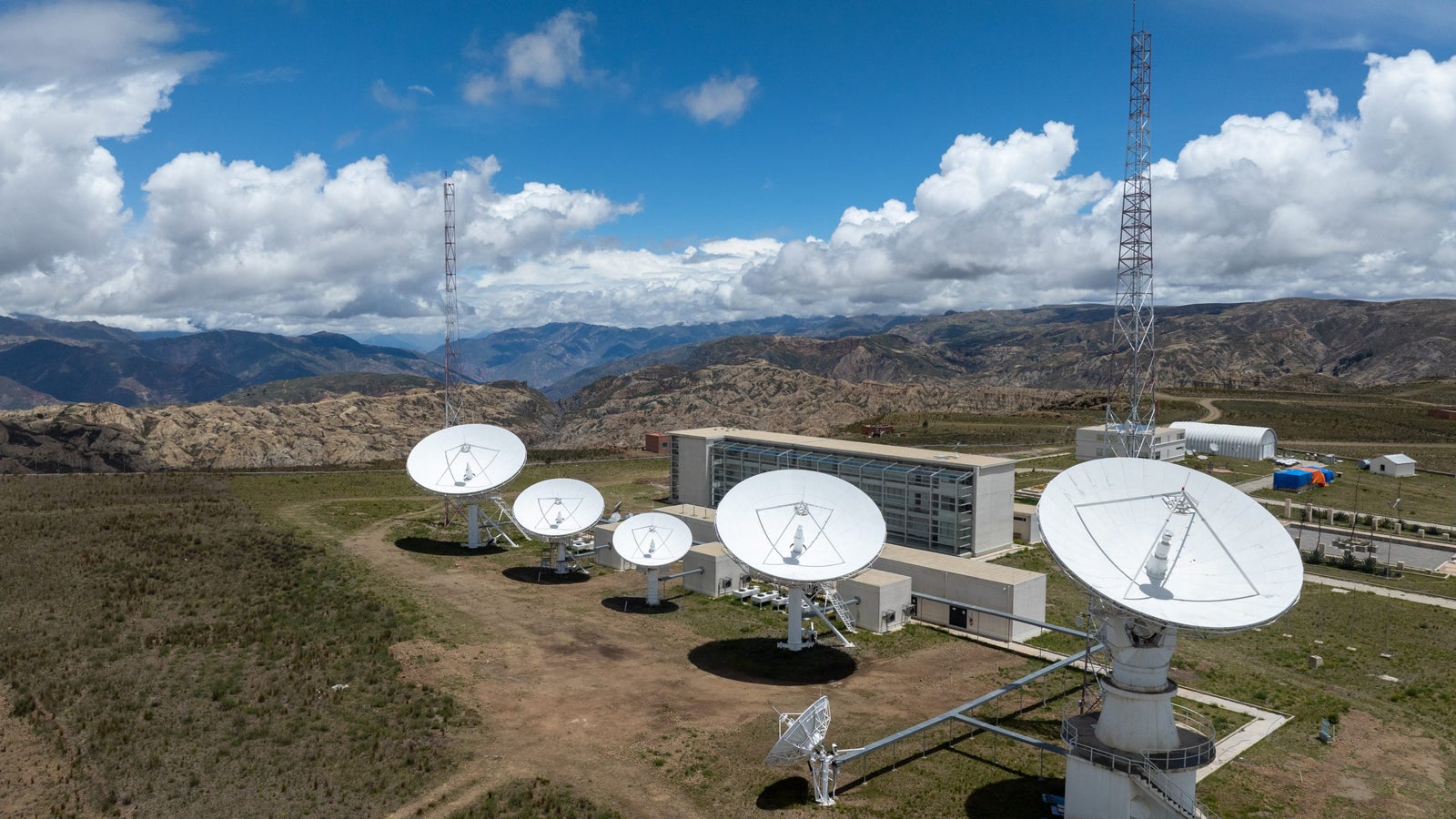 Aerial view of a satellite dish station with multiple large white satellite dishes on a grassy landscape, surrounded by mountains and under a partly cloudy sky. A modern building stands adjacent to the satellite dishes, with a tall communications tower in the background.