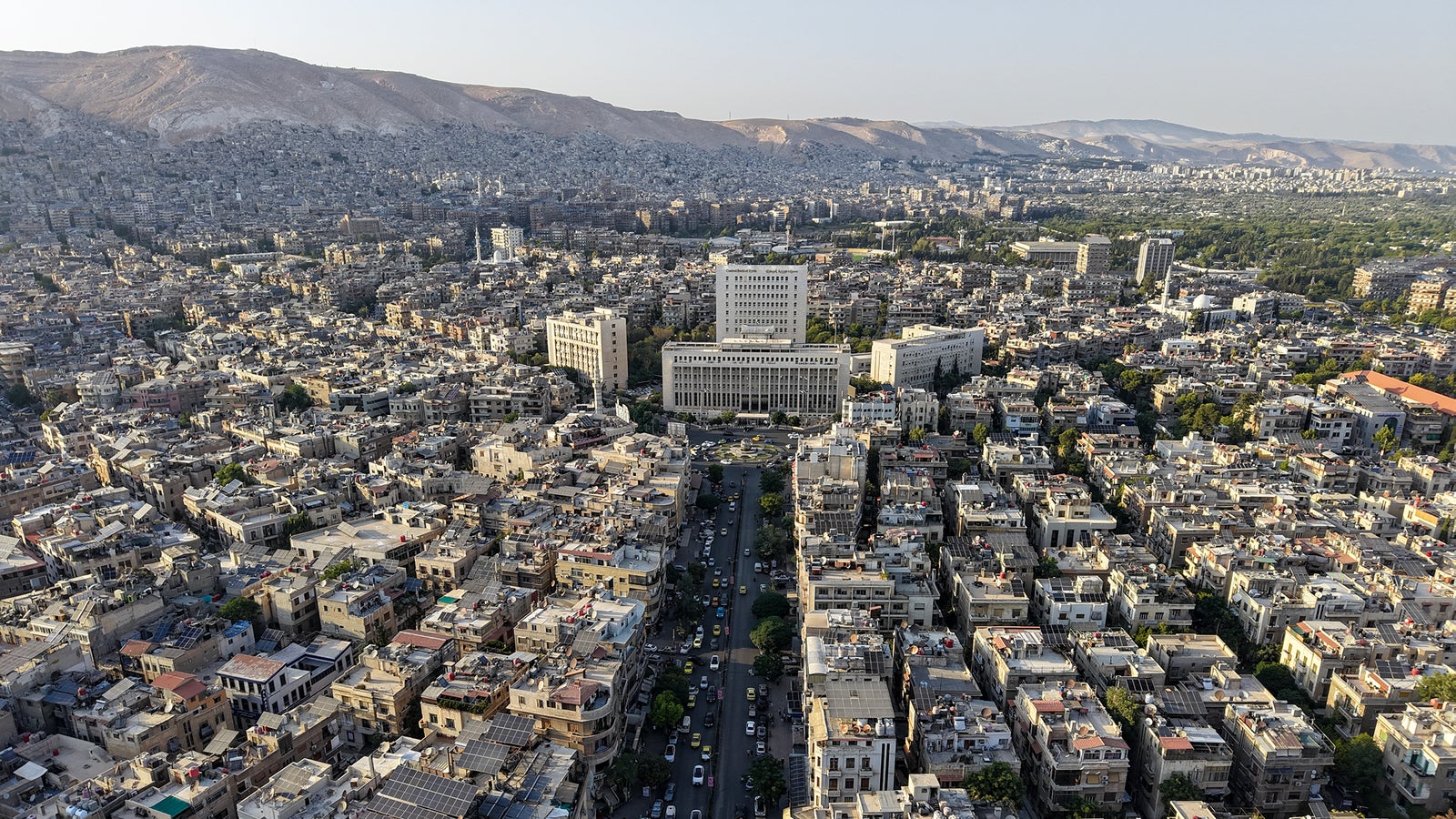 Aerial view of a densely populated urban area with a mix of low-rise buildings, some with solar panels on roofs, and a prominent modern structure in the center. The landscape is bordered by mountains, and several green trees are visible along a central road lined with cars.