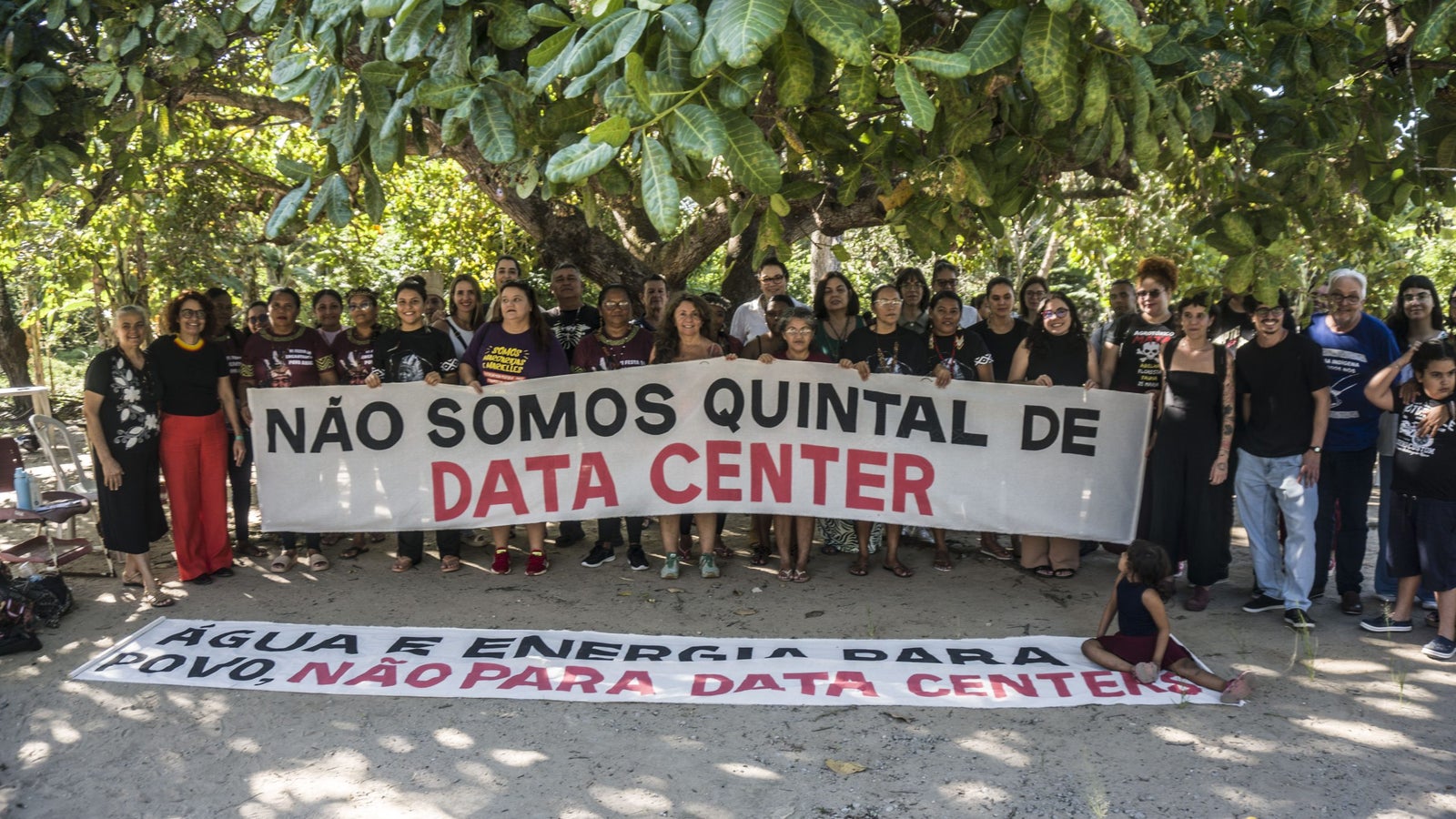 A group of people standing outdoors under a tree, holding banners that read "NÃO SOMOS QUINTAL DE DATA CENTER" and "ÁGUA E ENERGIA PARA POVO, NÃO PARA DATA CENTERS," expressing their opposition to data centers. The participants exhibit a range of ages and ethnicities, some dressed in black shirts, and they appear engaged and united.