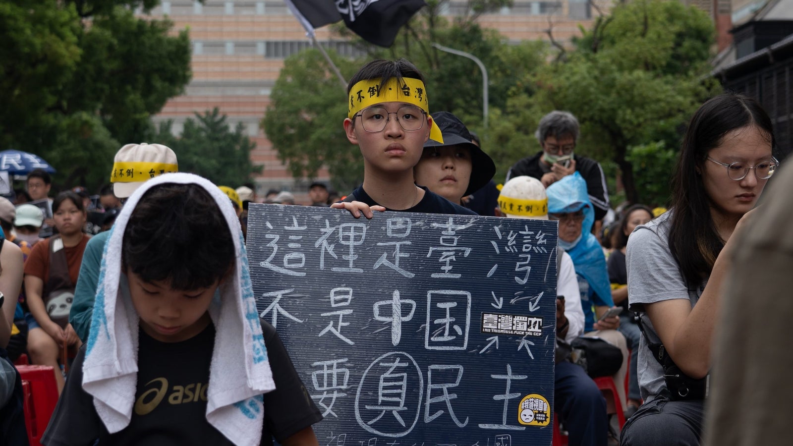 A photograph focused on a single protester holding a sign and wearing a yellow headband, amid a crowd of protesters seated in a street in Taiwan.