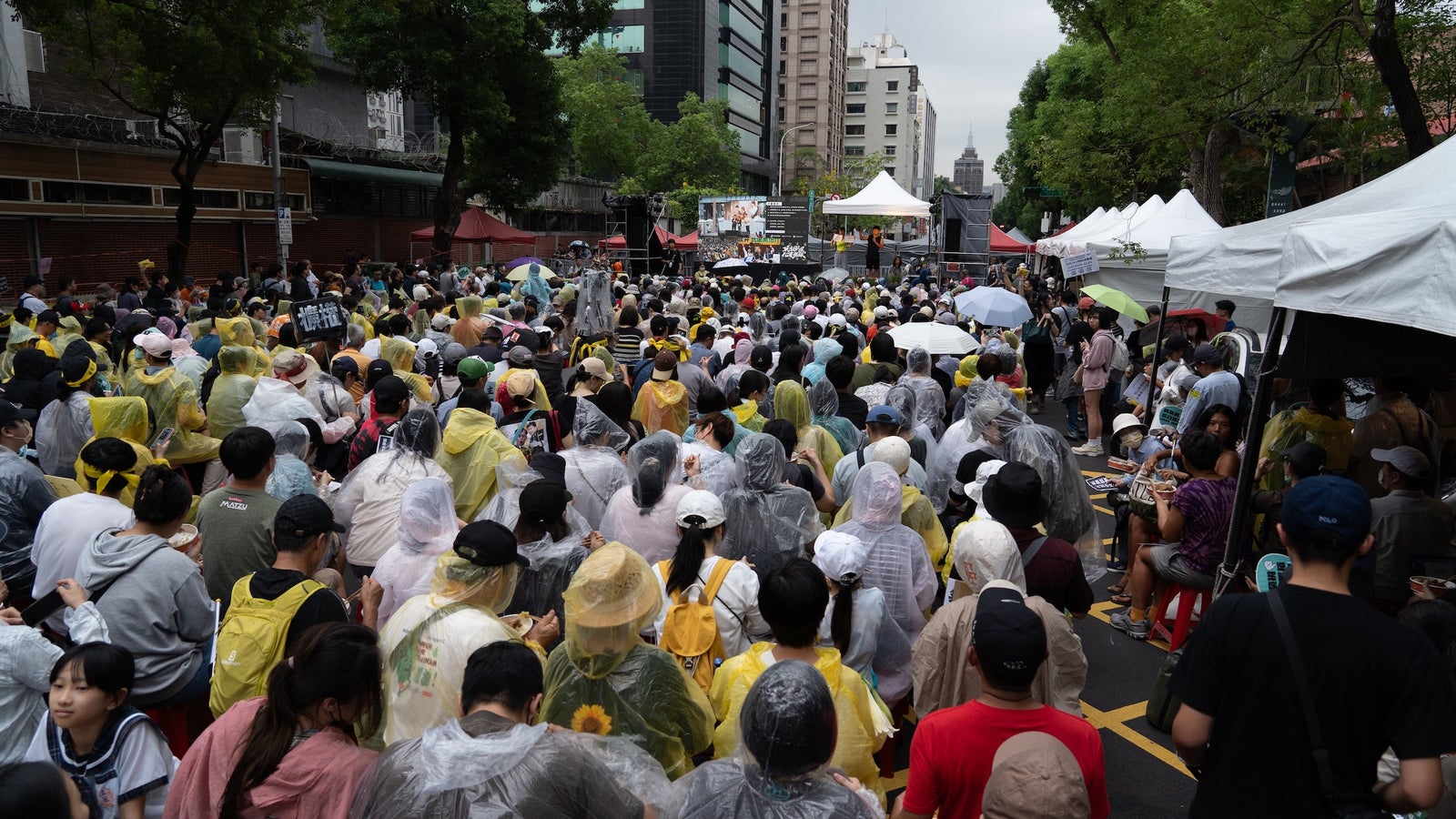 A photograph showing a large gathering of people, some wearing rain jackets or carrying umbrellas in a street in Taipei, Taiwan.