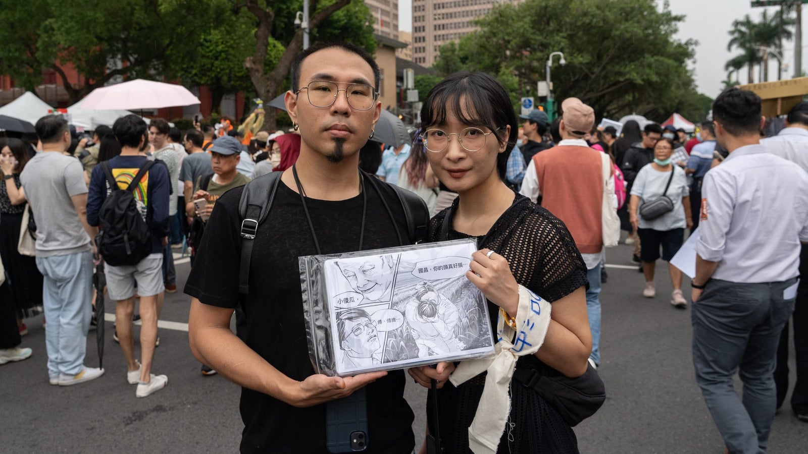 A photograph showing a man and a woman wearing black clothing and holding a cartoon drawing, standing in the street. Many people are walking and standing in the background behind them.