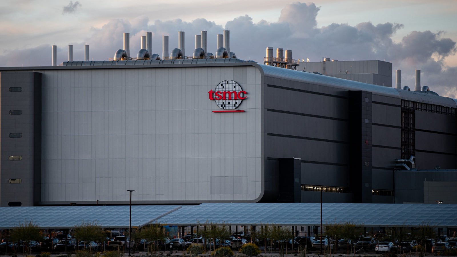 A photo showing a large silver building with the TSMC logo on the exterior of it and a large parking lot under solar panels.