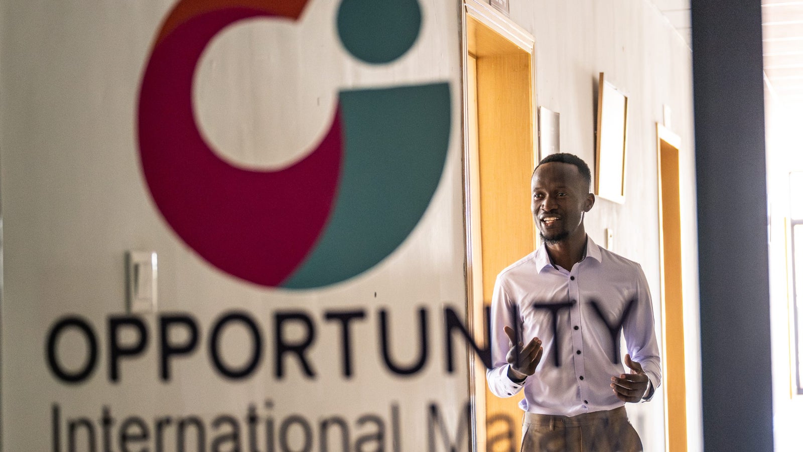 A man wearing a light purple shirt is standing in a corridor, speaking animatedly. In the foreground, a logo of "Opportunity International Malawi" in bold letters is visible.