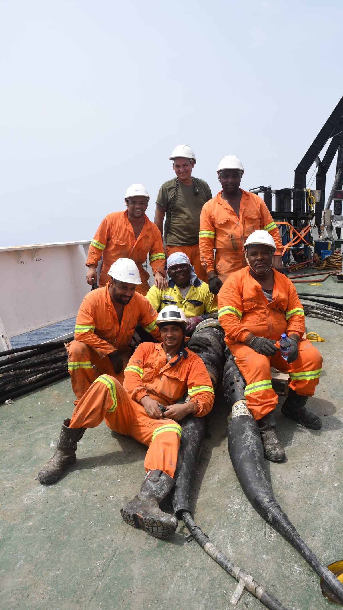 A group of seven men in orange work suits and white helmets, posing on a ship's deck with various cables and equipment visible in the background. They are smiling, with some sitting and others standing, set against a cloudy sky.