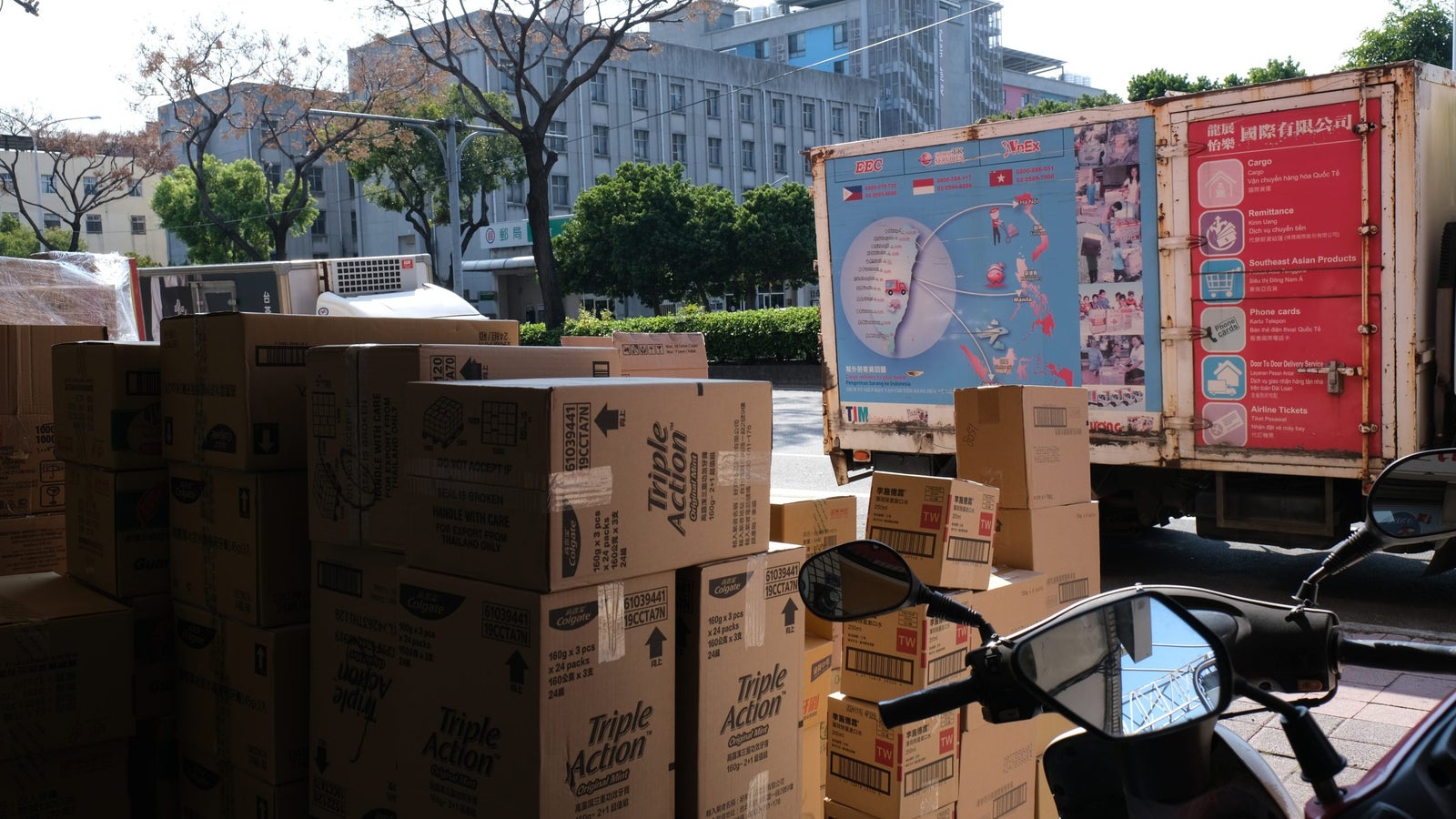 A stack of cardboard boxes labeled 'Triple Action' is piled in the foreground, with a motorcycle's handlebar and mirrors visible. In the background, a delivery truck with colorful advertisements is parked, alongside trees and buildings in a sunny urban setting.