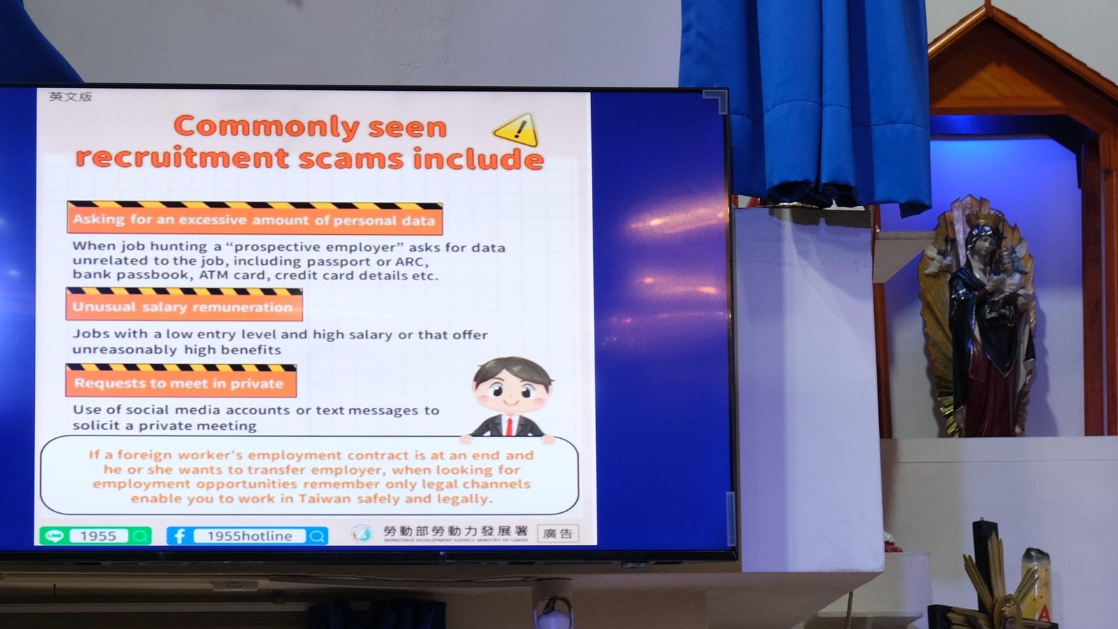 A presentation screen displaying information about commonly seen recruitment scams, including excessive personal data requests, unusual salary offers, and private meeting requests, alongside a cartoon character in business attire. The background features a blue curtain, religious statues, and a visible television setup.