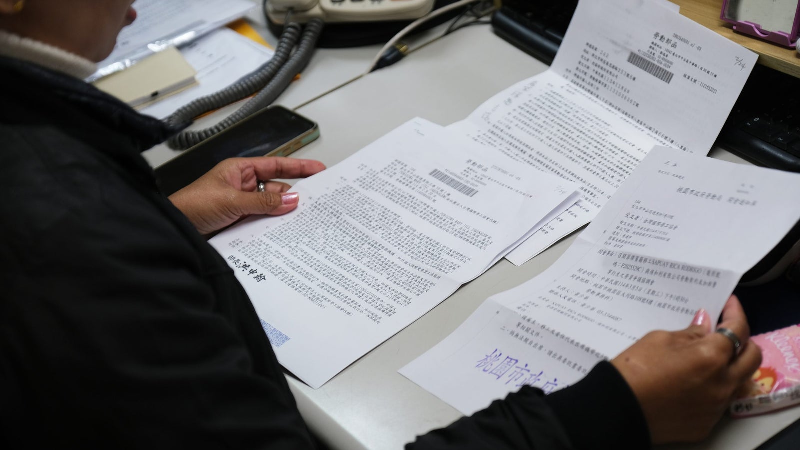 A person is seated at a desk holding a document, surrounded by several papers, a telephone, and a smartphone, with text visible on the documents, indicating a work or study environment.