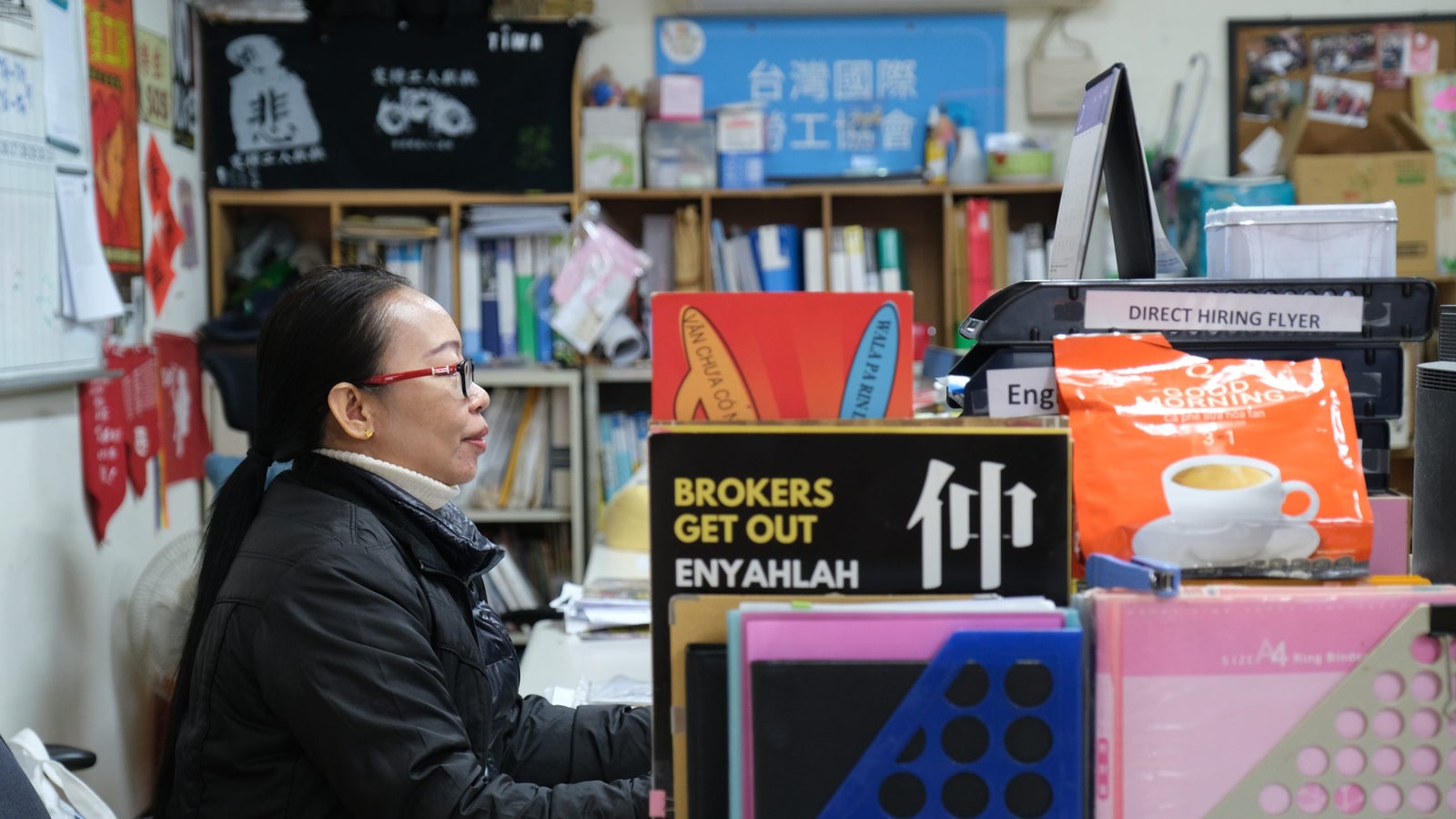 A woman with long hair tied back sits at a desk, working on a computer. She wears a black jacket and glasses. The desk is cluttered with various items including a direct hiring flyer, colorful binders, and a package of instant coffee. Behind her, shelves filled with books and materials are visible, along with posters and notes on the walls.