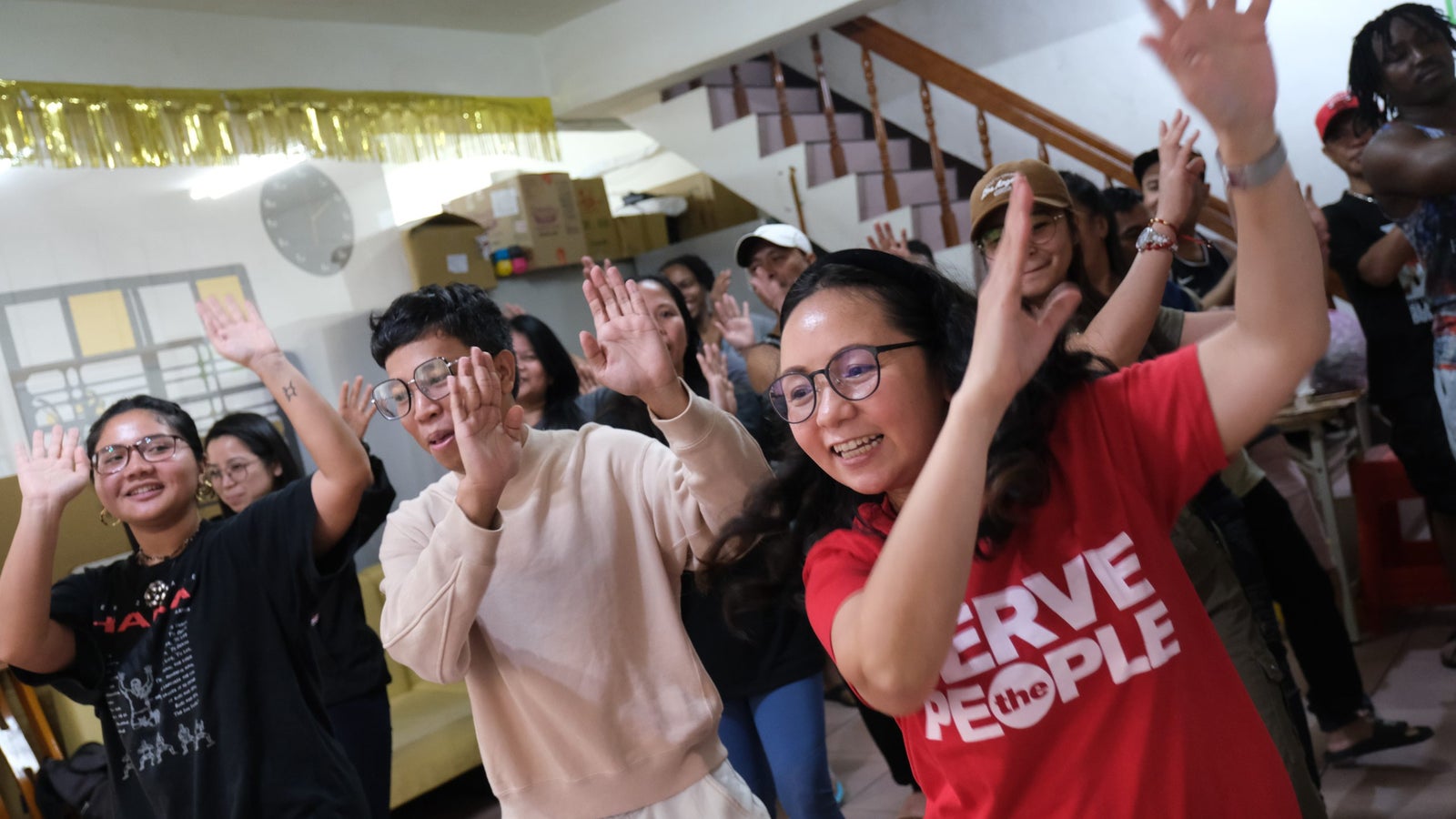 A diverse group of people participating in a lively indoor dance activity, with hands raised and smiling faces. The participants include individuals of different backgrounds wearing casual clothes, and a festive atmosphere is suggested by decorations in the background.