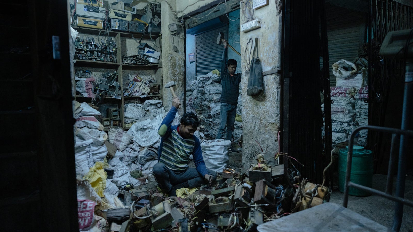 Two men working in a cluttered, dimly lit room filled with piles of discarded electronics and bags. One man crouches on the floor, holding a hammer, while the other stands in the background with a tool raised, amidst shelves packed with various electronic components and equipment.