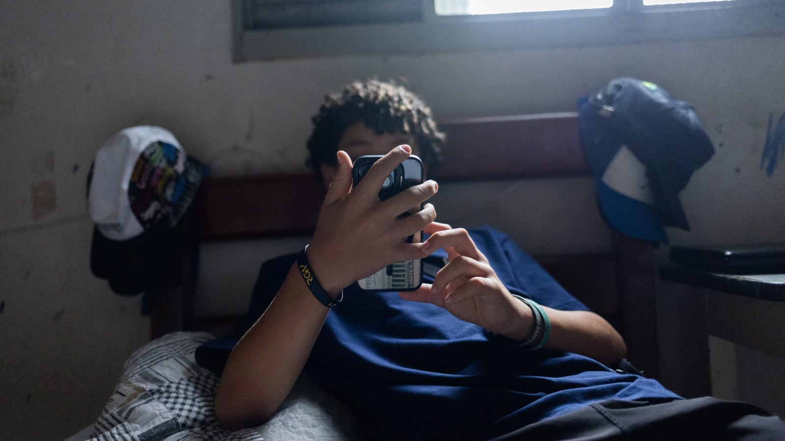 A person with curly hair lying on a bed, holding a smartphone and looking at its screen, with a window letting in soft light and various baseball caps hanging on the wall behind them.