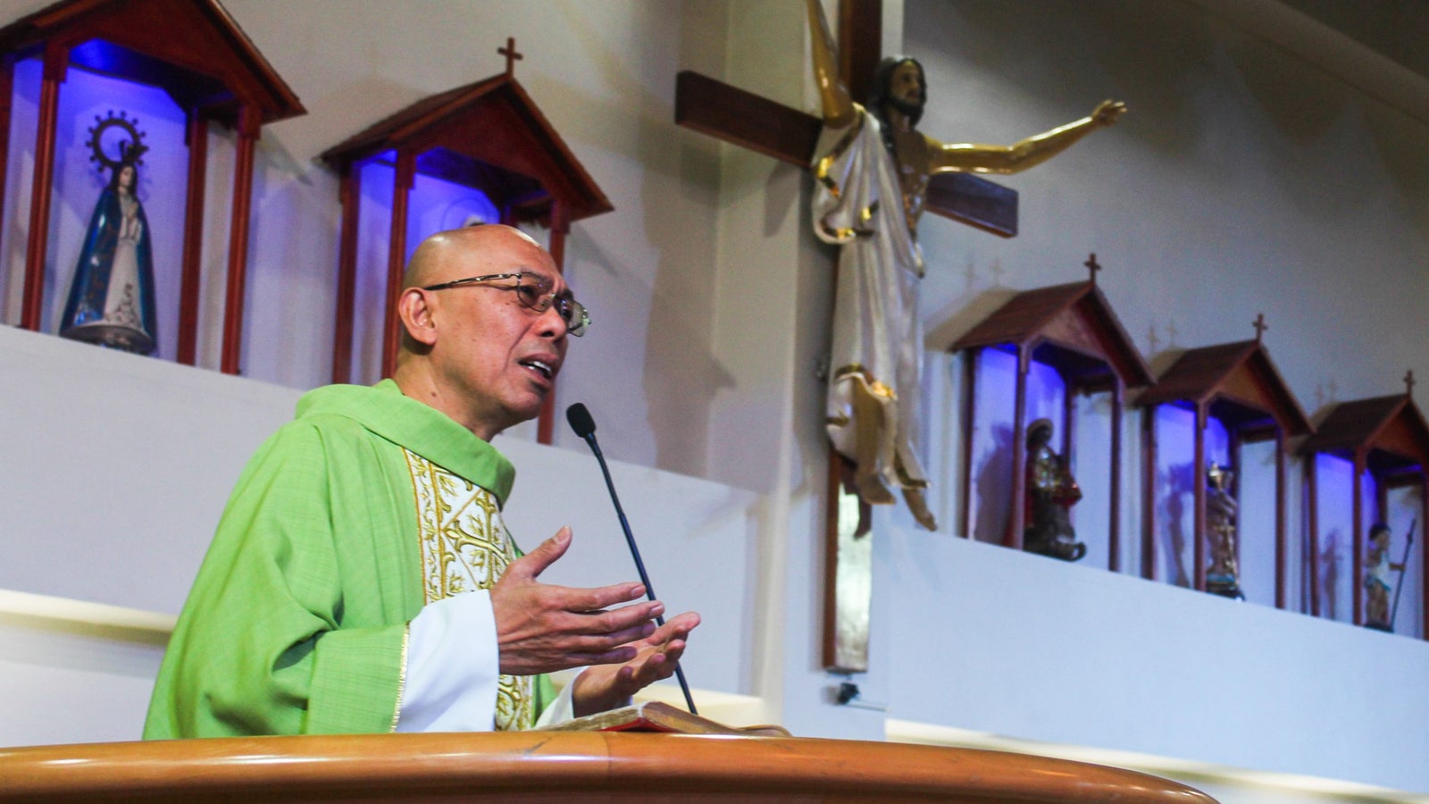 A priest in a green vestment is speaking at a podium in a church, with a figure of Jesus on a cross and several small religious statues displayed behind him.