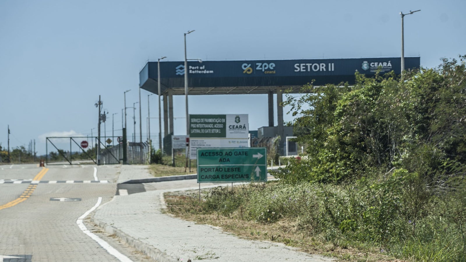 Entrance to a logistics area, featuring a large sign indicating 'Port of Rotterdam' and 'Sector II', with directional signs for access to different gates and loading areas, surrounded by greenery and a clear blue sky.