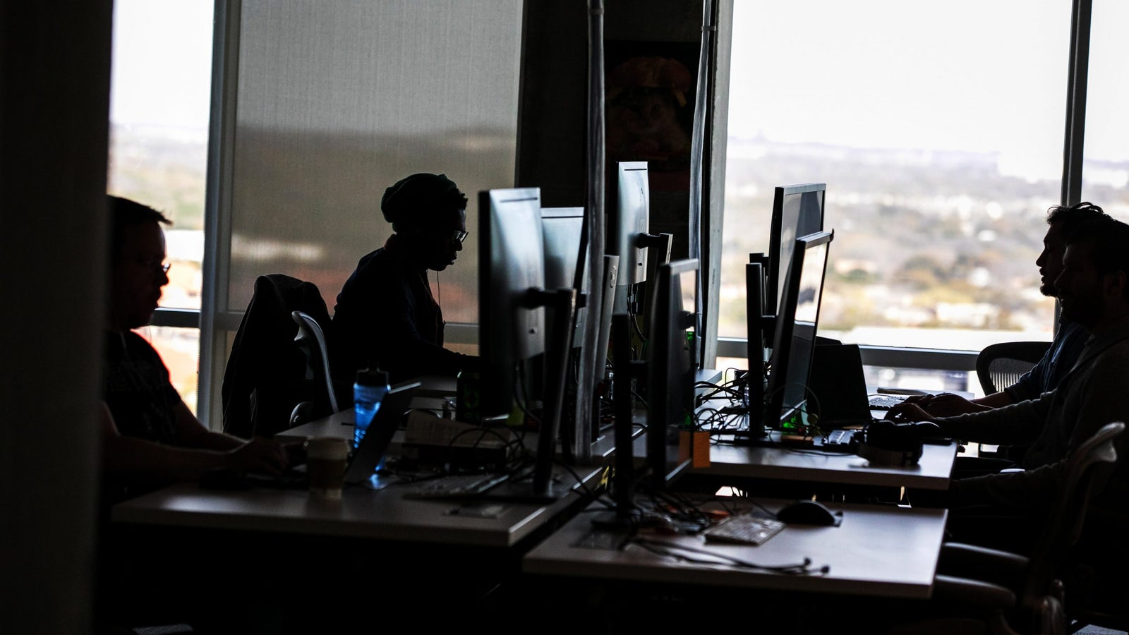 Silhouettes of several people sitting at desks with computers in a bright office space, illuminated by natural light from large windows, creating a contrast against the darker interior.
