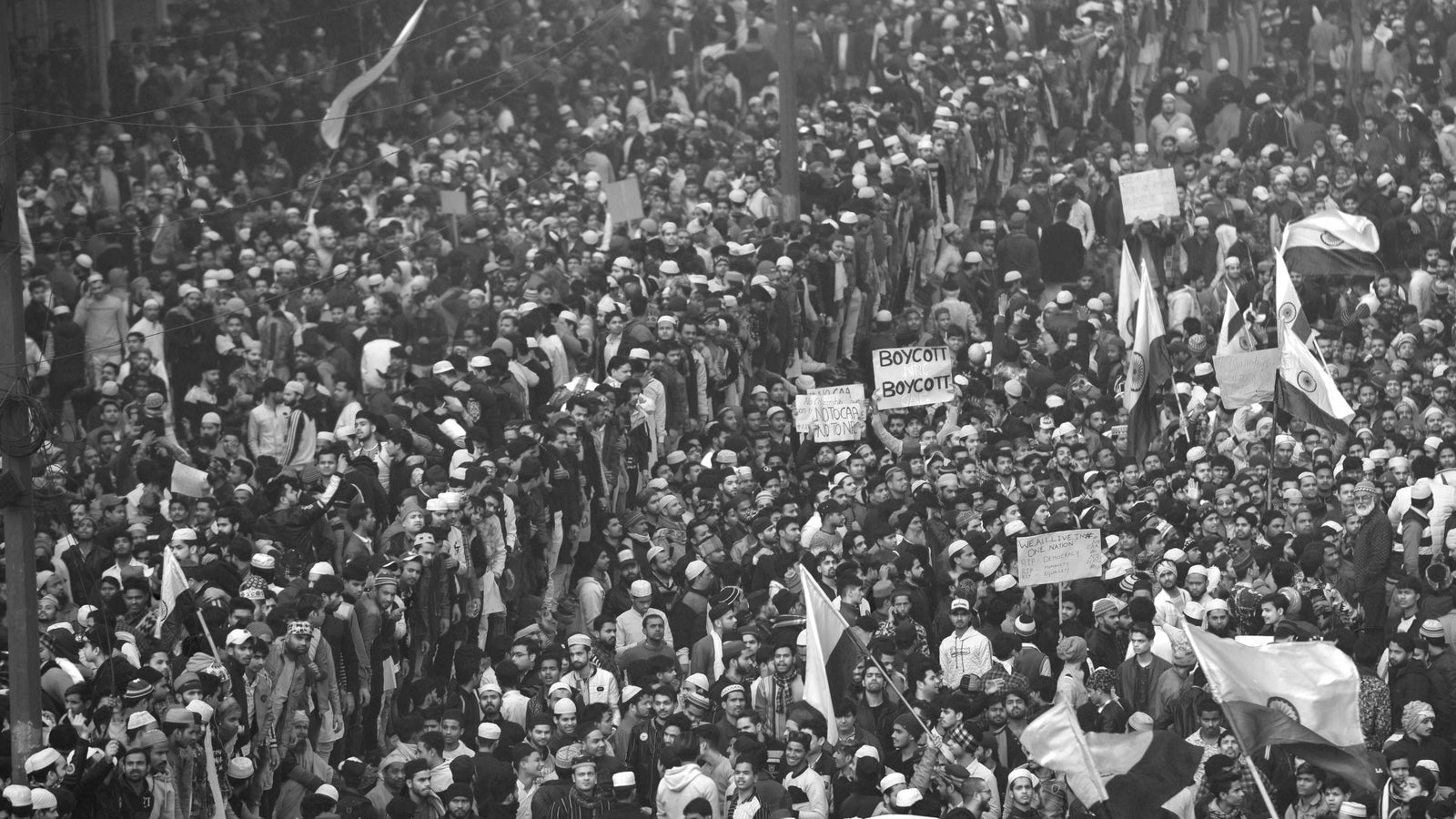A black and white photograph showing a massive crowd of people protests on the street of an Indian city, many are waving white flags and holding signs.