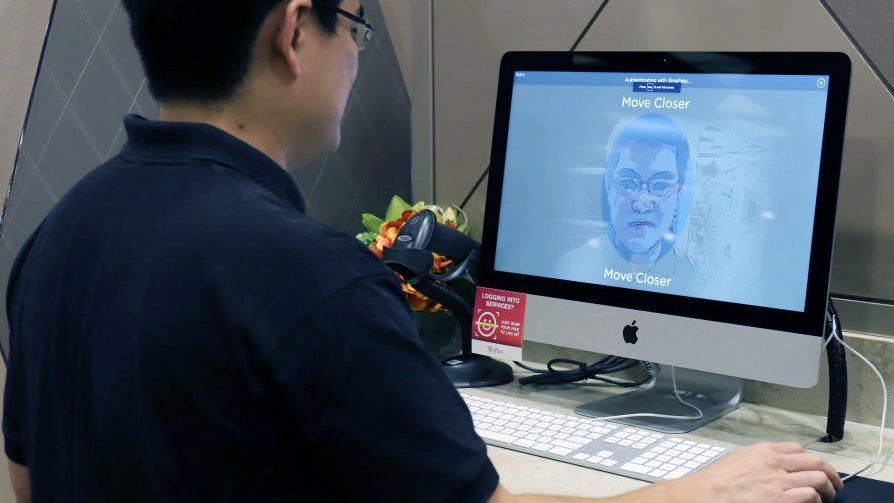 A person stands in front of an iMac computer displaying a face recognition interface that prompts them to 'Move Closer.' The setting features a decorative plant on the desk beside the computer.