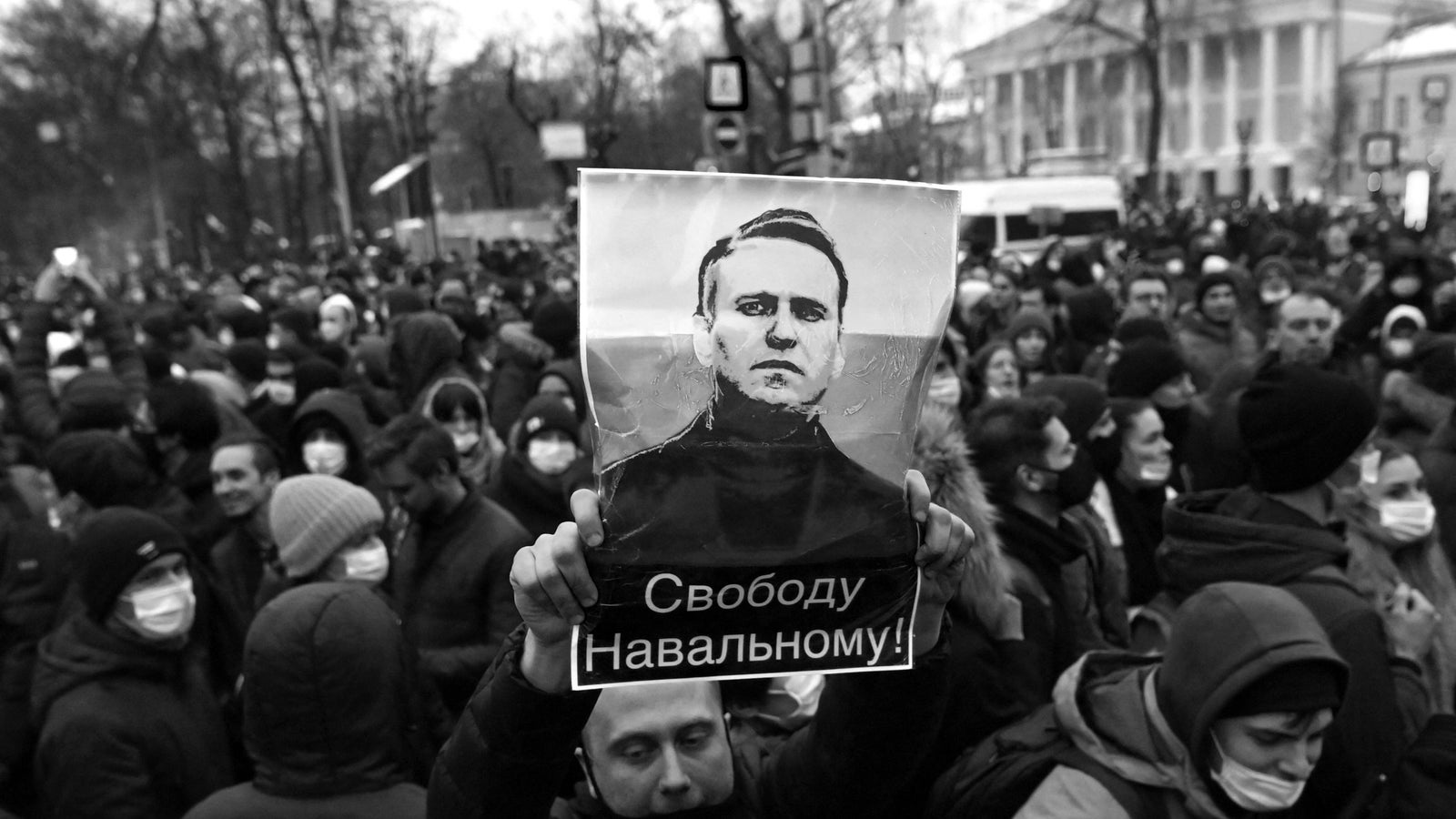 A black and white photograph showing a large group of people, the person in the center is holding up a picture of Alexei Navalny with Russian text under it.