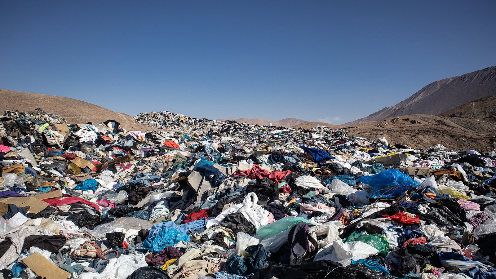 A vast landfill covered with heaps of discarded clothing and plastic debris under a clear blue sky.