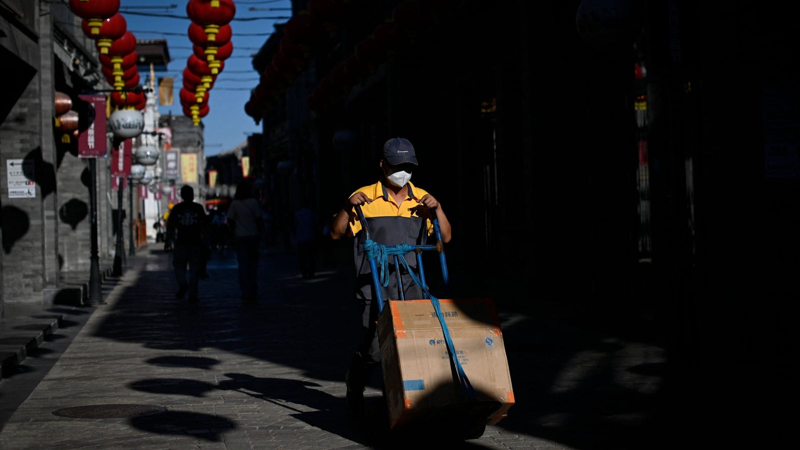 A worker wearing a face mask and a yellow and gray uniform pushes a cart with large cardboard boxes through a dimly lit street decorated with red lanterns, casting long shadows on the cobblestone pavement.