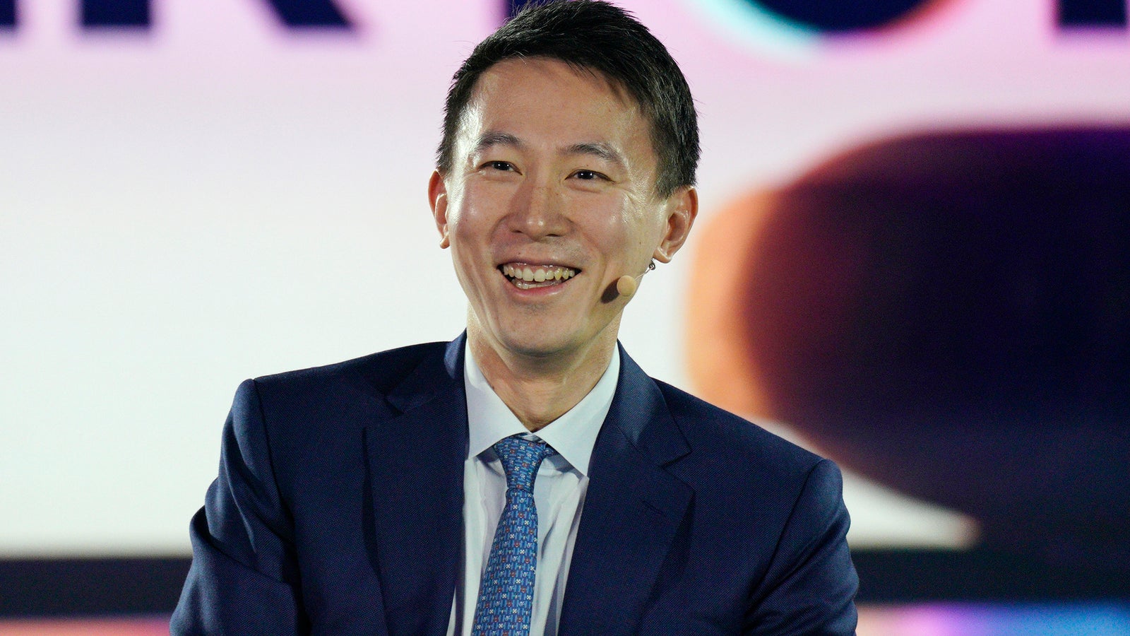 A smiling man in a dark suit and tie, speaking at an event with a colorful background behind him.