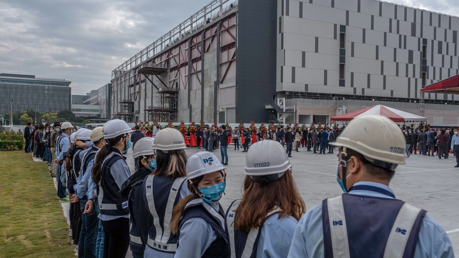 A photograph showing rows of people wearing hardhats standing outside a large factory on a cloudy day.