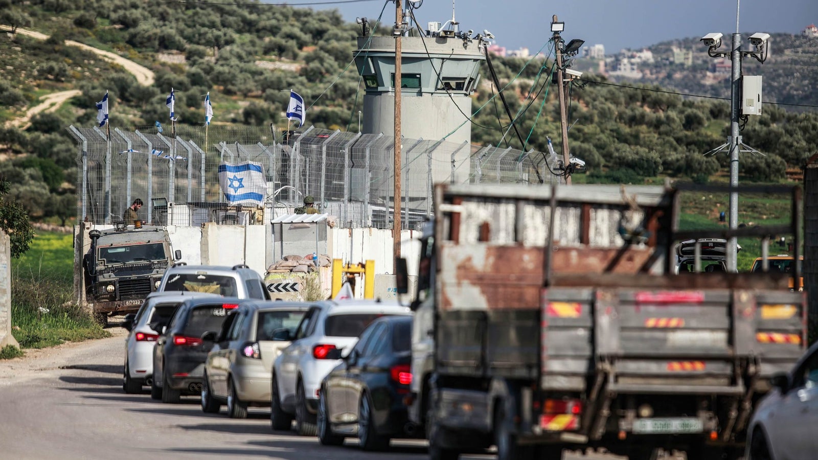 A line of vehicles waits on a road near a security checkpoint surrounded by fencing and surveillance equipment, with a watchtower and Israeli flags visible in the background.