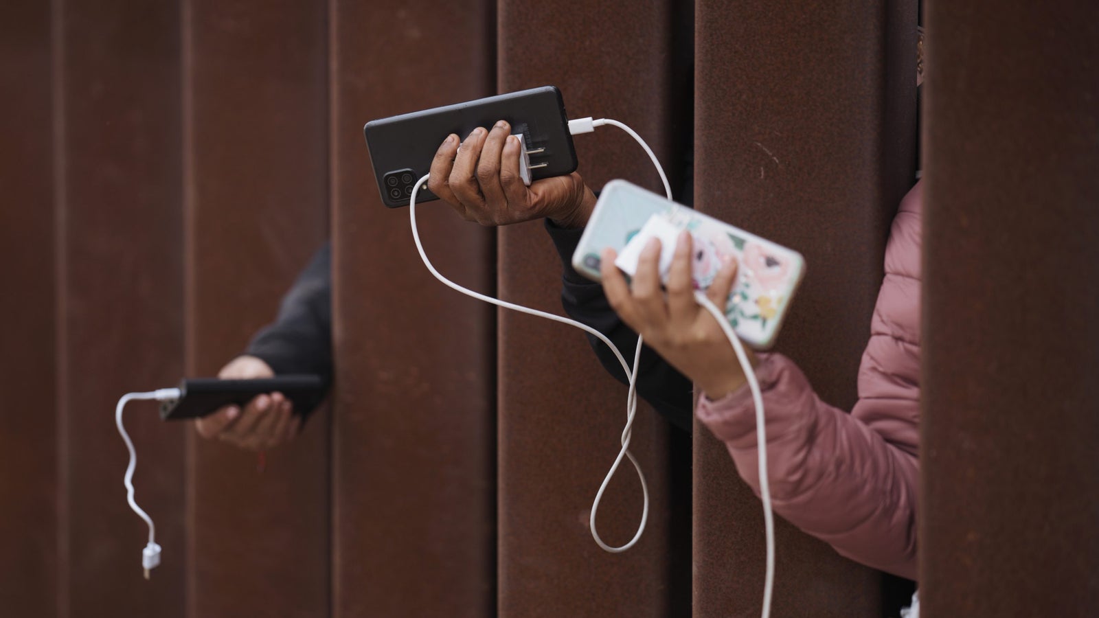 Two hands holding smartphones with charging cables visible, positioned between vertical metal bars.