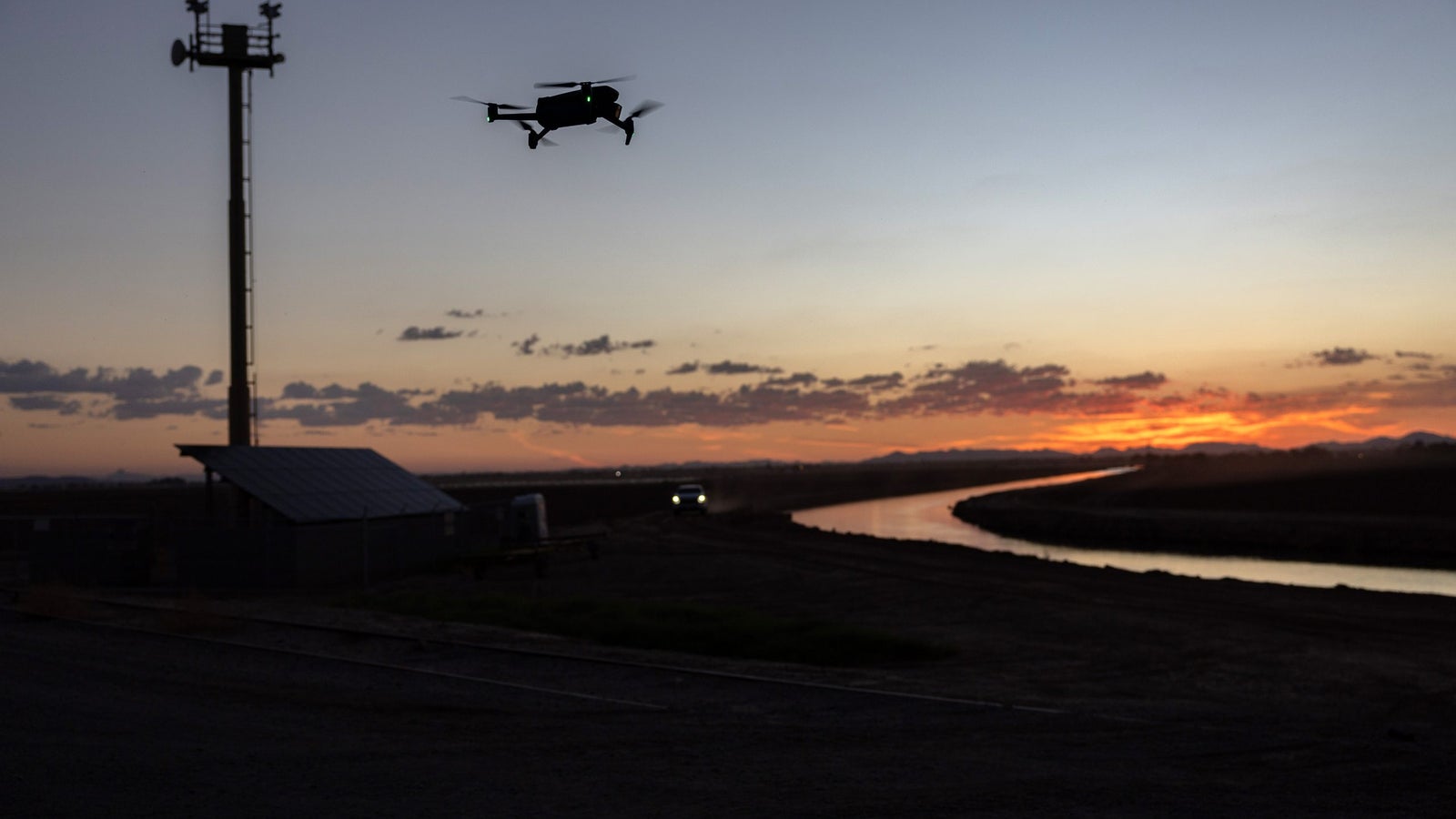 A drone flies in the foreground against a colorful sunset, with a silhouetted communication tower and a meandering waterway in the background.
