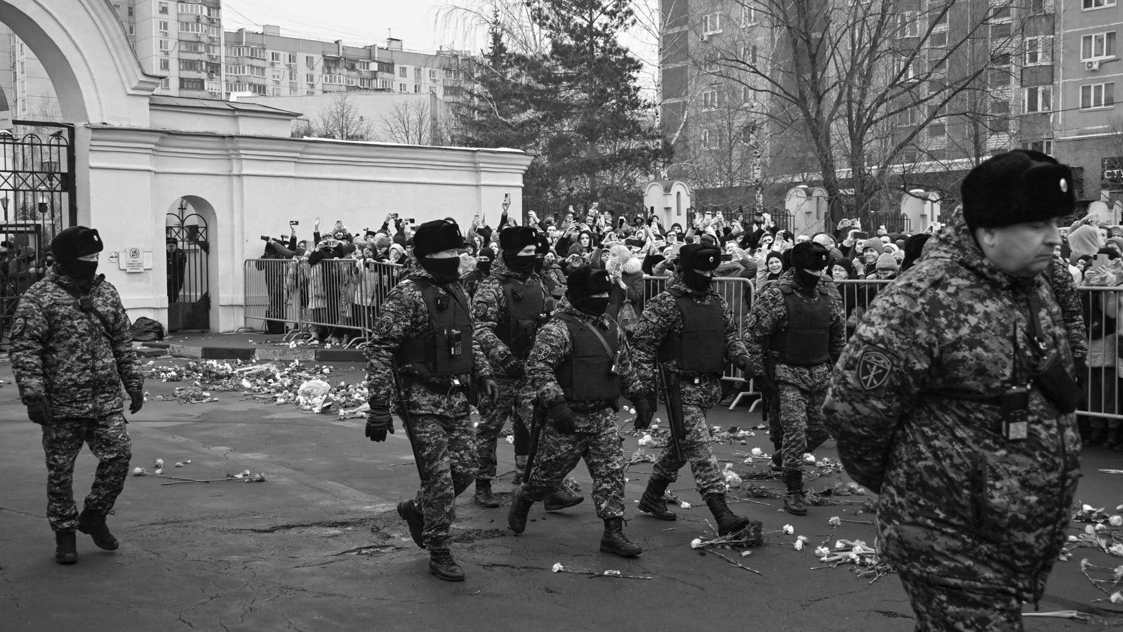 A black and white photograph showing a number of security officers wearing camouflage and body armor, walking along a street past a large crowd.