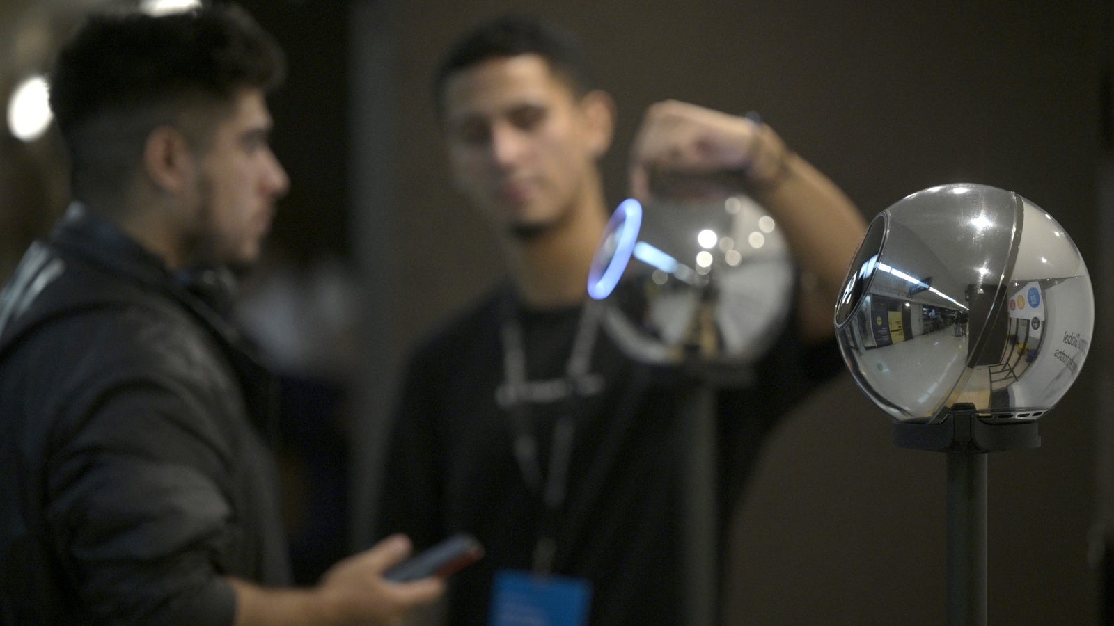 A photograph showing a Worldcoin orb in the foreground and a person helping another person scan their face with an orb in the background.