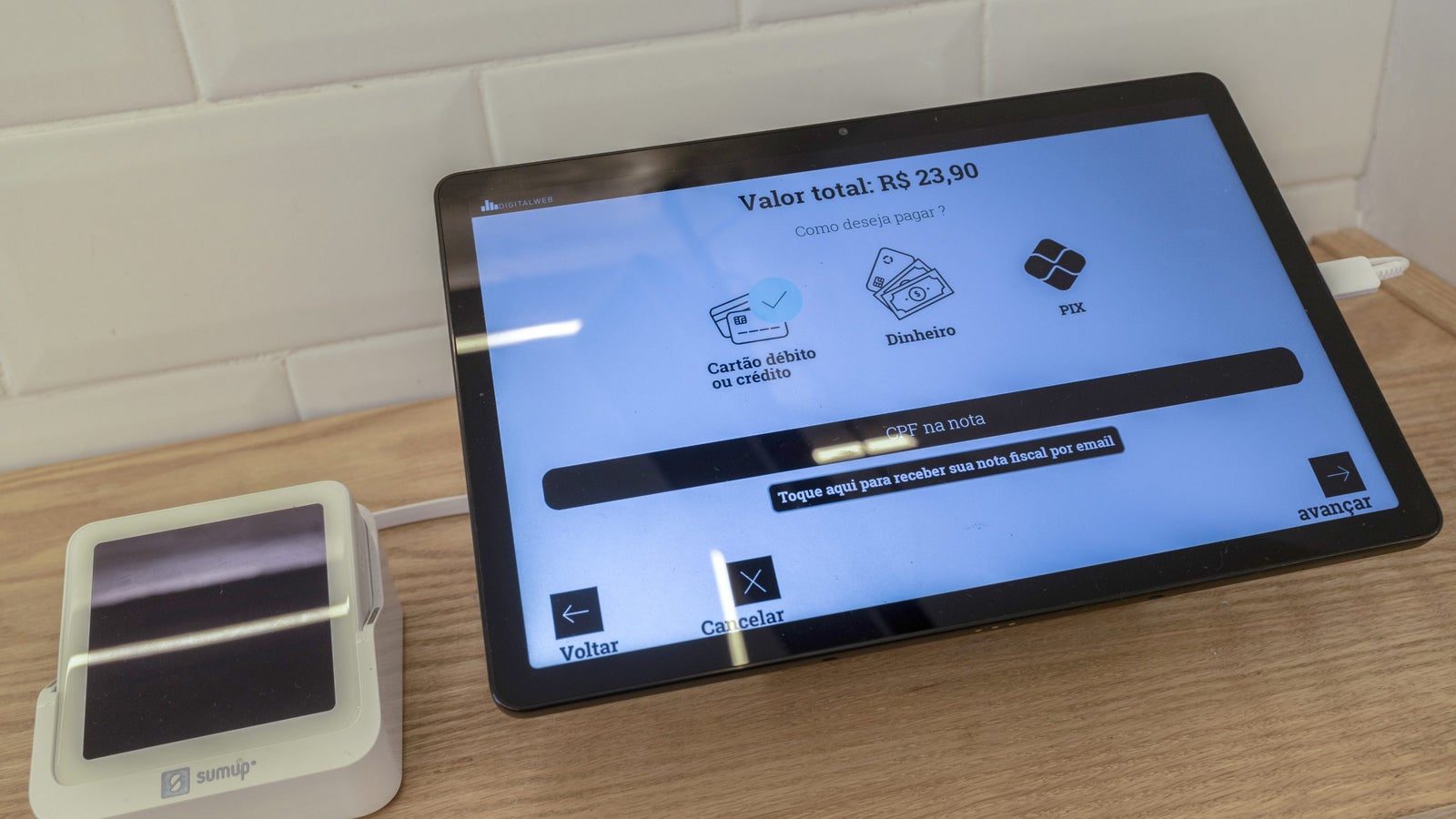 A touchscreen payment terminal displaying a total amount of R$ 23,90, with options for payment methods including debit/credit card, cash, and PIX, along with buttons for returning or canceling the transaction, and an option for receiving a digital receipt via email.