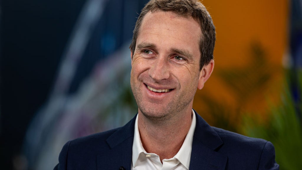 Man with short brown hair and a slight beard, smiling while wearing a dark suit and white shirt, seated in front of a blurred background with hints of greenery and colorful graphic elements.