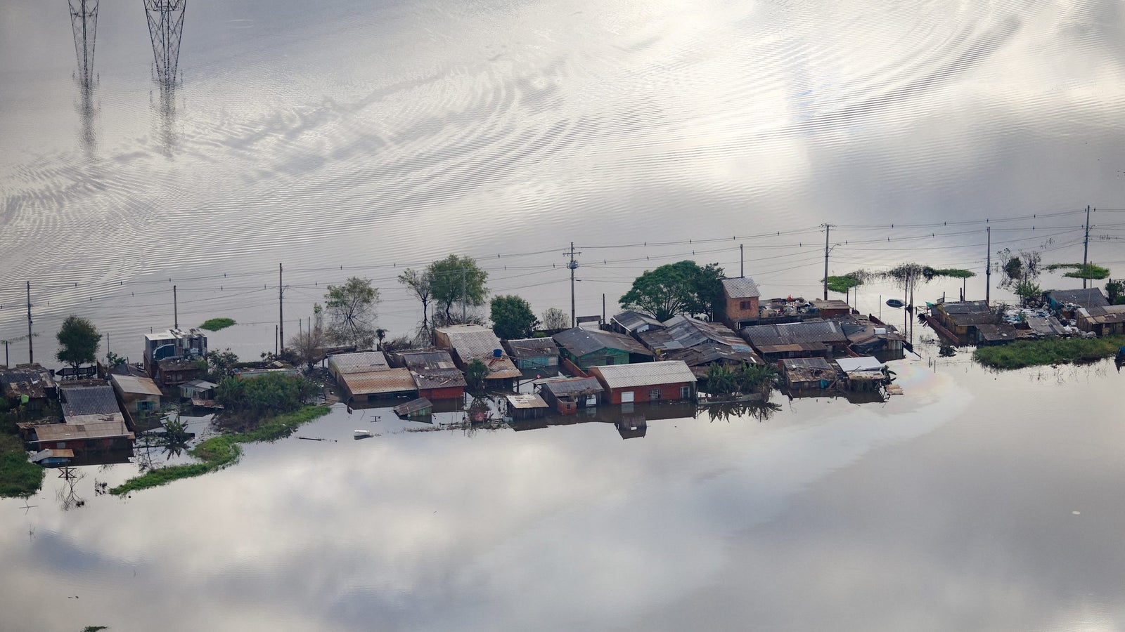 An aerial view of a flooded area showing several small houses partially submerged in water, with power lines in the background and reflections on the water's surface.
