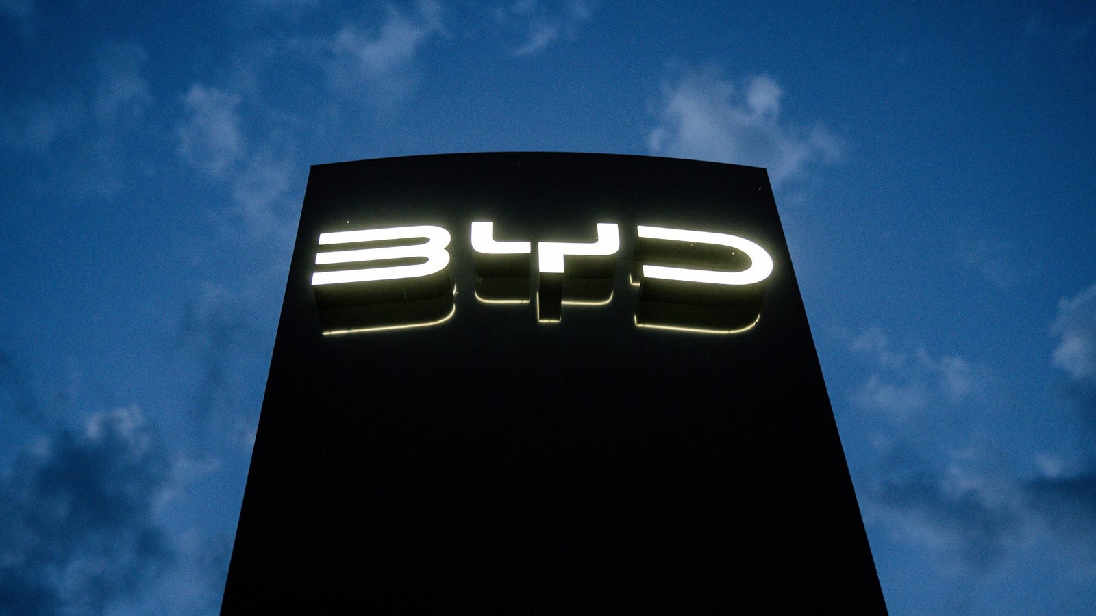 Illuminated BYD logo on a dark background, with a blue sky and clouds above.
