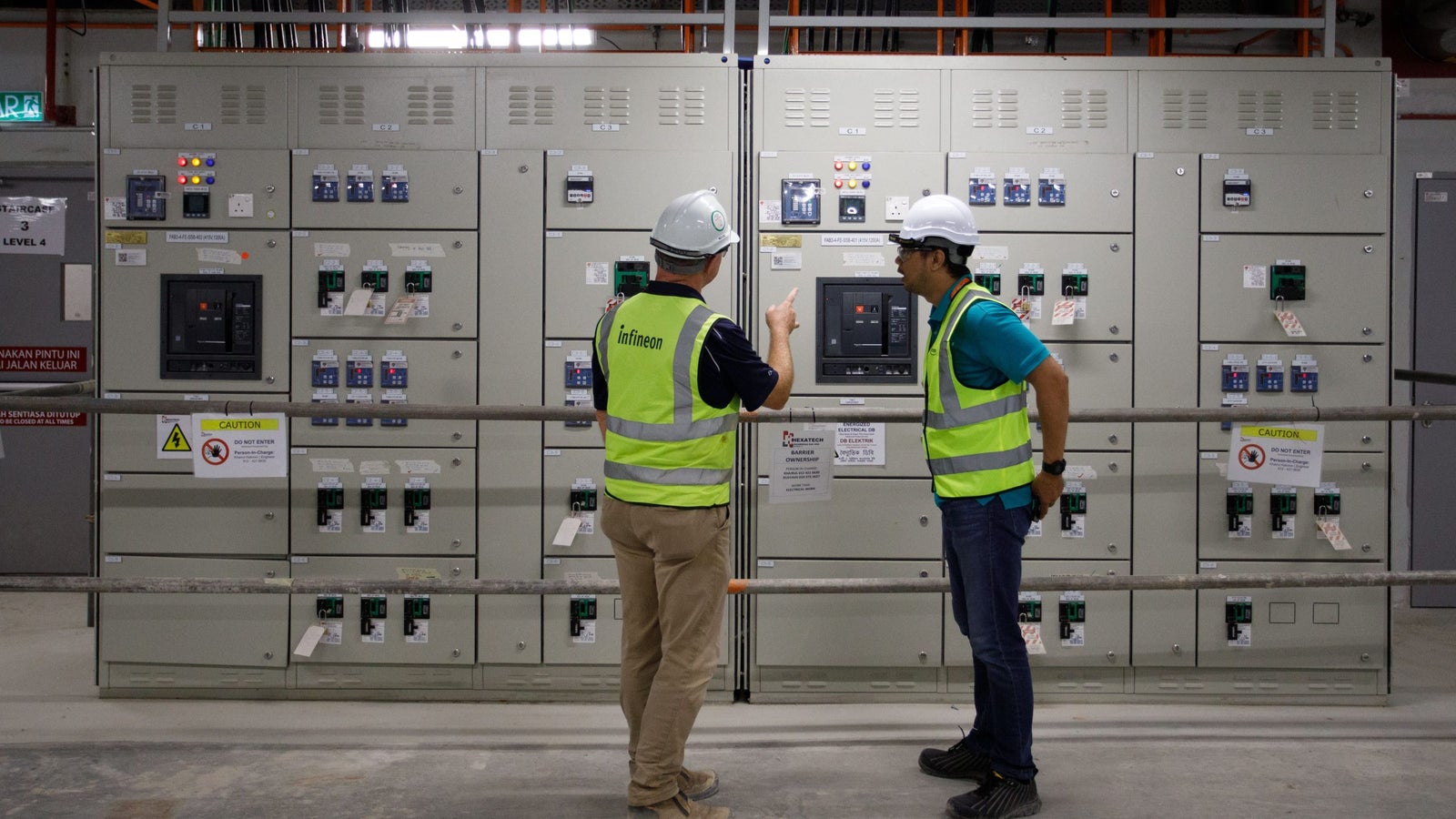Two men in safety vests and hard hats are discussing equipment in front of a panel of electrical control boxes with various switches and indicators.