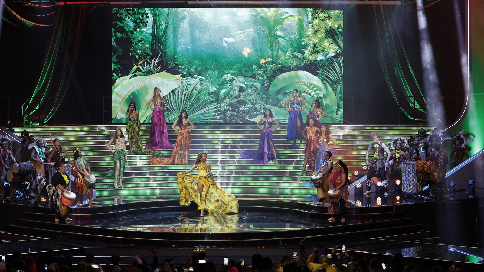 A photograph showing a number of women wearing colorful gowns on a large stage, surrounded by muscians with drums, in front of an audience, many holding cell phones in their hands.